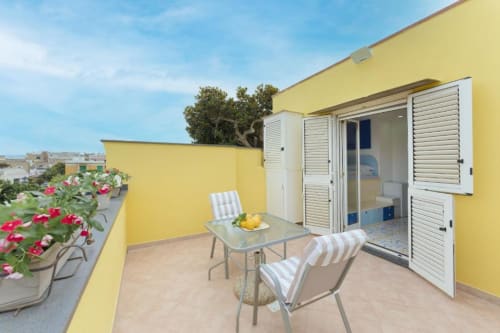 Terrazza soleggiata di IL POETA a Procida con lettini a righe, tavolo, piante fiorite e ingresso in giallo con blu