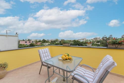Terrazza gialla di IL POETA a Procida con tavolo da pranzo, sedie a righe e vista sulla città con edifici colorati sotto il cielo blu