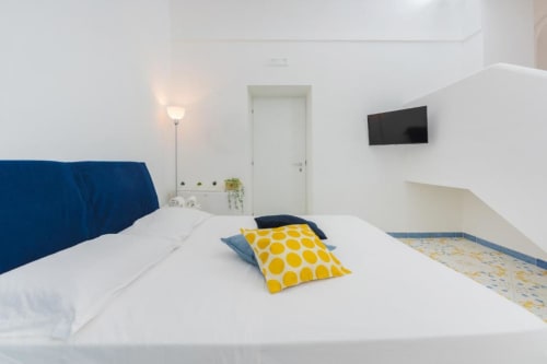 Camera moderna a IL POETA a Procida con testata blu, letto bianco, cuscino con pois gialli, TV a muro e arredamento bianco