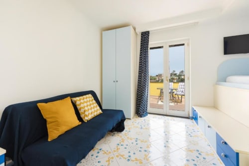 Camera moderna e luminosa a IL POETA a Procida con divano blu, accenti gialli, accesso alla terrazza e vista sulla città