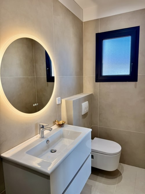 Salle de bain moderne de la villa Figari avec miroir ovale illuminé, lavabo blanc, toilettes et fenêtre aux encadrements sombres