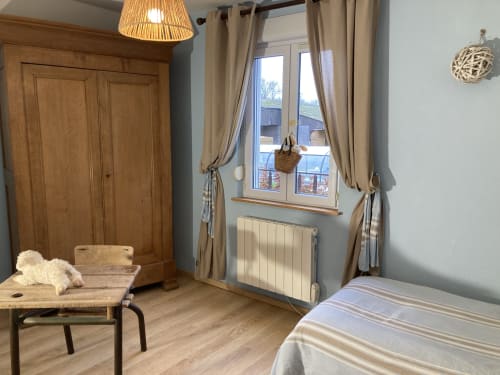 Chambre lumineuse avec armoire en bois, grande fenêtre vue sur le lac, rideaux beiges, radiateur et lit confortable
