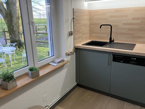 Cuisine de cottage moderne avec dosseret en bois, robinet noir et grande fenêtre donnant sur les jardins verdoyants du Domaine De La Ferme