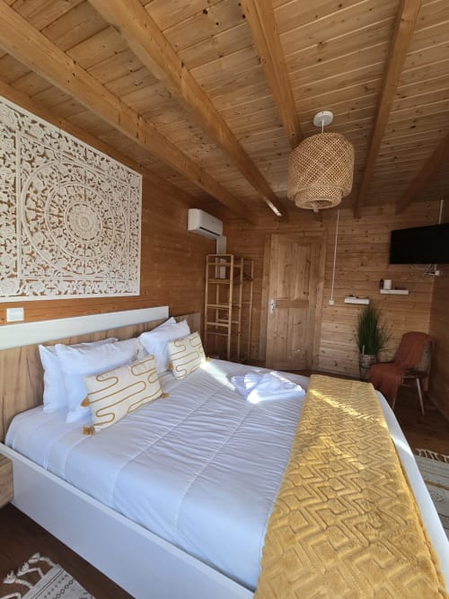 Mountain Wood Village B bedroom with wooden beams, queen bed with gold pillows, ornate white wall art, woven pendant light,