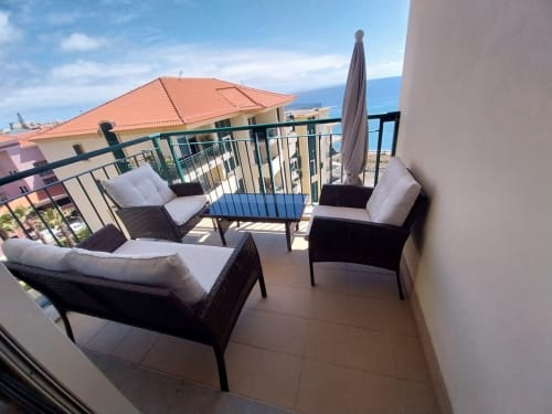 Ampla varanda da Casa Delores em Funchal com vista para o mar, móveis de vime, guarda-chuva e edifícios com telhados de terracota