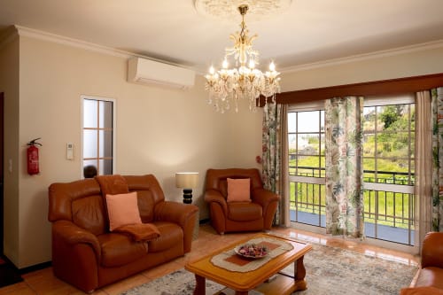 Elegante sala de estar da Paradise House Calheta com assentos em couro conhaque, lustre e vista para o jardim