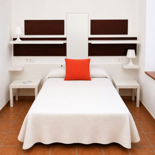Modern bedroom with white bed, orange pillow, dark wood headboard, white nightstands, and terracotta tile flooring at