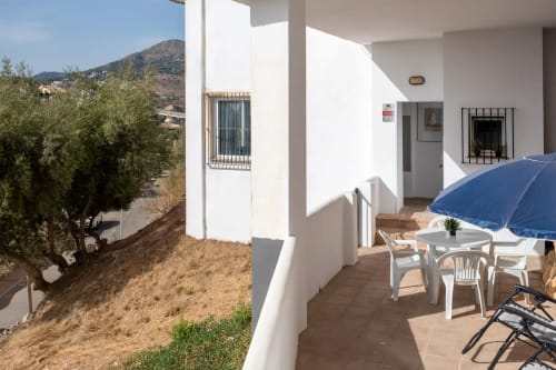 Sunny terrace of apartment in Málaga with blue umbrella, seating area, and mountain views with olive trees