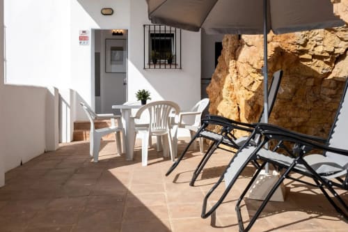 Sunny terrace with white and black lounge chairs, dining table, rock wall backdrop at Apartamentos Turísticos Axarent, Málaga