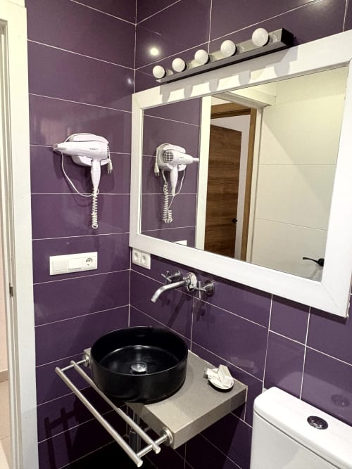 Modern bathroom with purple tiled walls, black round sink, white-framed mirror, wall-mounted hair dryers, and toilet at