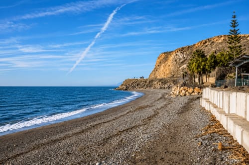 Pebble beach at Almayate Alto with turquoise sea, white protective barriers, rocky cliffs and palm trees under blue sky