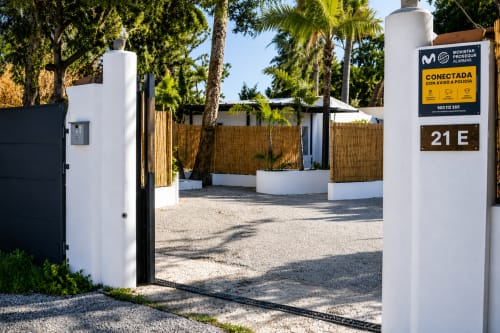 Modern white apartment entrance with wooden gates at Axarent Almayate Alto, featuring palm trees and gravel courtyard,
