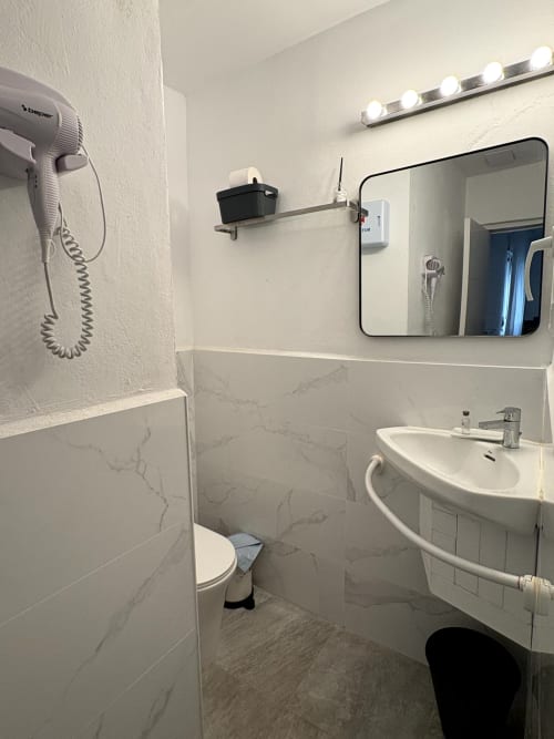 Modern bathroom with marble tiles, round sink, mirror with vanity lights, toilet, and wall-mounted hair dryer at Apartamentos