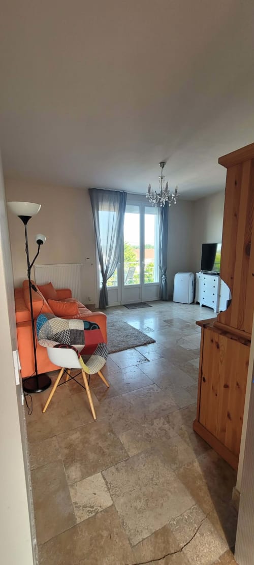 Suite lumineuse avec balcon à Villa Varco, Auxonne. Canapé orange, mobilier en bois, TV et vue sur la ville par portes vitrées.