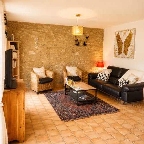 Salon rustique aux murs en pierre, fauteuils en osier, canapé en cuir noir et carrelage en terre cuite du Domaine Bellavigne Gîtes