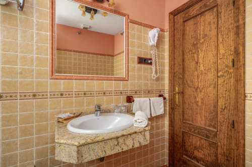 Salle de bain de la suite Balcón De Los Montés avec carrelage beige, lavabo en granit, porte en bois et touches de corail.