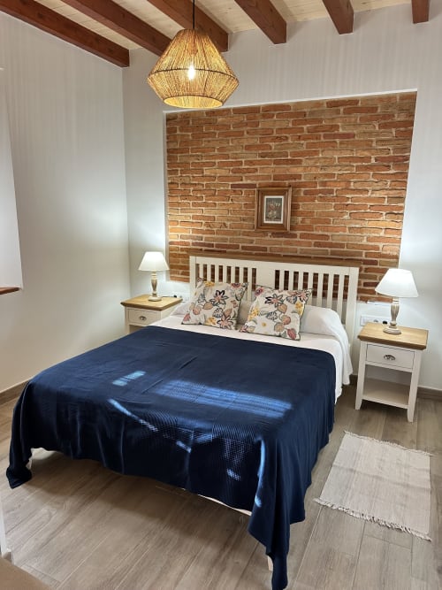 Dormitorio rústico en El Escondite De Pedro Malillo con pared de ladrillo visto, cama blanca, manta azul marino, lámpara colgante y elementos de madera