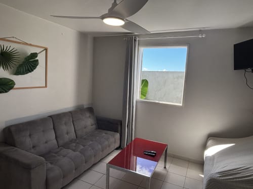 Modern cottage living room with gray sofa, red table, ceiling fan, TV, and window overlooking pool at Domaine De Marly,