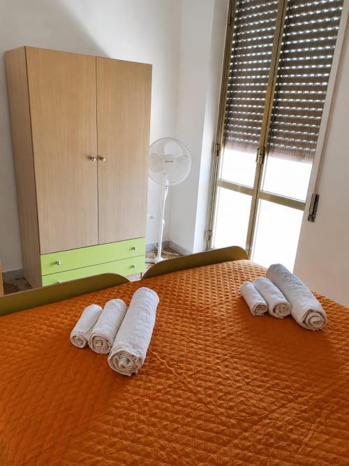 Camera da letto di Villa Leuka San Lorenzo a Noto con copriletto quilted arancione, asciugamani bianchi arrotolati, armadio in legno, ventilatore, e