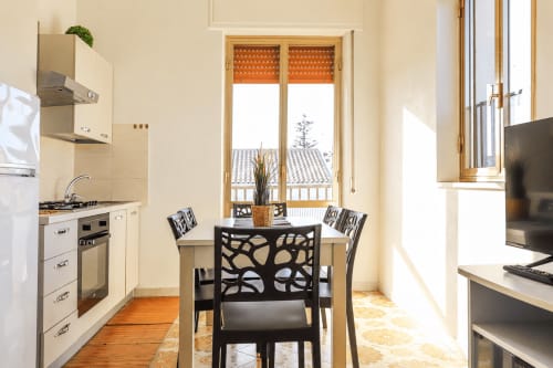 Zona pranzo moderna di Villa Leuka San Lorenzo, Noto, con sedie nere intorno a tavolo rotondo, cucina a vista e porte-finestre