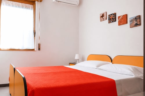 Camera luminosa di Villa Leuka San Lorenzo a Noto con letto arancione, coperta rossa, cuscini bianchi e vista sul giardino