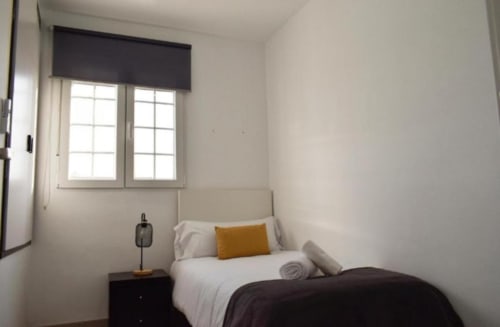 Dormitorio moderno y luminoso en Gestyp-AT de Mérida con ropa de cama blanca, almohada mostaza, mesita oscura y gran ventana con vistas a la ciudad