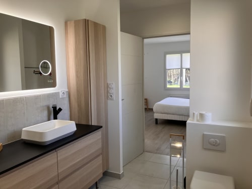 Salle de bain moderne à Domaine Des Bois Argentés avec meuble en bois, lavabo blanc, miroir éclairant et vue sur le jardin