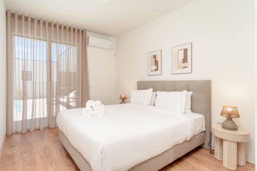 Bright modern bedroom with beige upholstered bed, white linens, pool view balcony access, and contemporary furnishings at