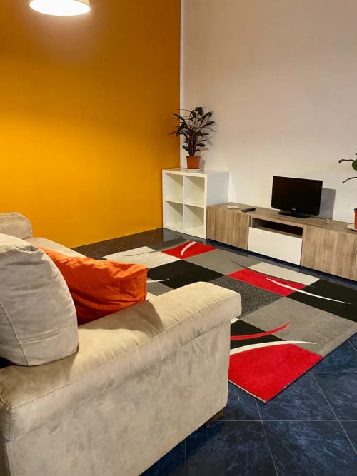 Elegante soggiorno della B&B Fossatello a Genova con divano color crema, TV, tappeto geometrico, scaffali bianchi e parete gialla