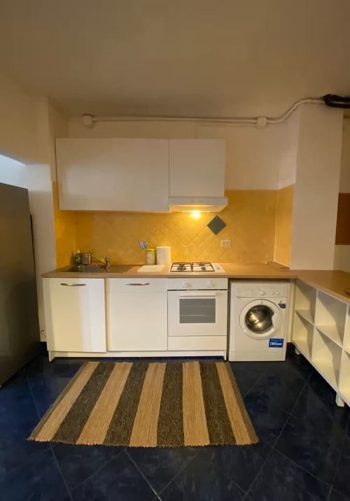 Cucina moderna nell'appartamento di Genova con mobili bianchi, piastrelle gialle, piano cottura a gas, lavatrice e tappeto rigato