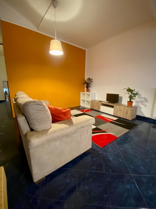 Soggiorno moderno della struttura B&B Fossatello Genova con divano crema, parete accent arancione, tappeto geometrico, TV e pianta in vaso