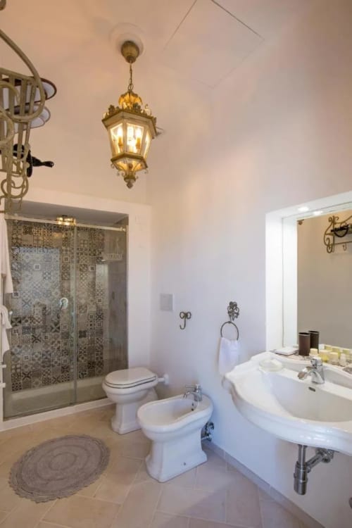 Elegante bagno al Relais Palazzo Cacace con pavimenti in marmo, lampada a sospensione in ottone, doppi lavabi e cabina doccia decorata