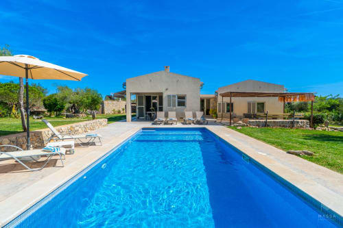 Villa de lujo en Artà con piscina rectangular, tumbonas, sombrilla y casas de piedra bajo cielo azul despejado