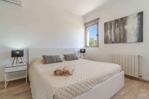 Dormitorio moderno con cama blanca, almohadas a rayas, suelos de madera, vista a la ventana y decoración minimalista en Artà, España