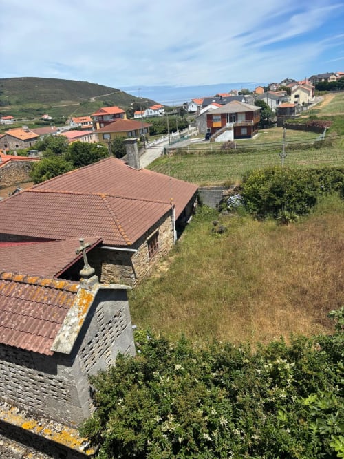 Vista del jardín del Hotel O Esqueiro en Malpica, A Coruña, con casas de piedra tradicionales, tejados de tejas rojas y loma verde
