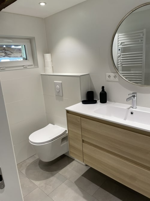 Modern bathroom in Ferme Freysz B&B with white tile walls, wall-mounted toilet, wooden vanity, round mirror, and towel heater