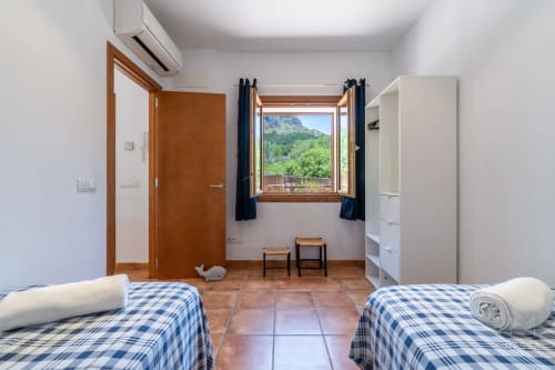 Dormitorio luminoso con dos camas individuales, vistas a la montaña desde ventana de madera, almacenamiento blanco y suelo de terracota en villa de Artá