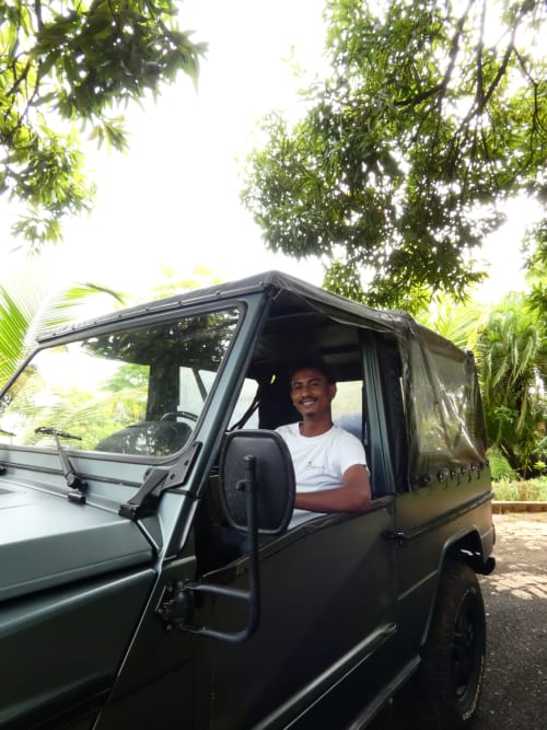 Homme souriant dans un SUV vert à la Belamandy Lodge, Nosy Bé, sous des arbres tropicaux