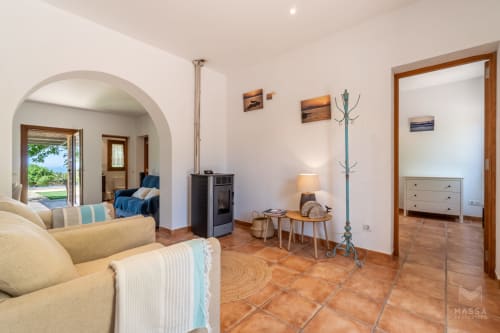 Luminoso salón de la villa Can Jaumí en Artá con sofá color crema, suelo de barro, vigas de madera, chimenea y vistas al mar