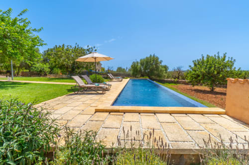 Lujoso oasis de piscina en Massa Properties Artà con tumbonas, sombrilla y jardines exuberantes bajo cielo despejado