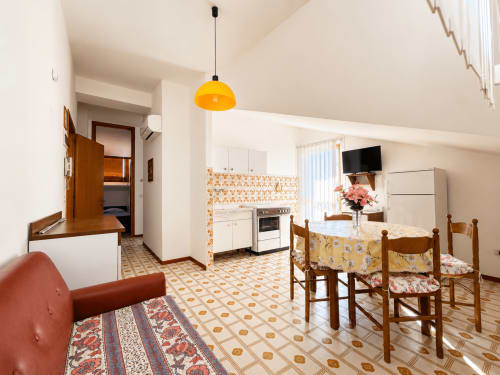 Luminosa zona living open-plan all'hotel Anthea di Lignano Sabbiadoro con cucina, tavolo da pranzo, divano rosso e lampada a sospensione gialla