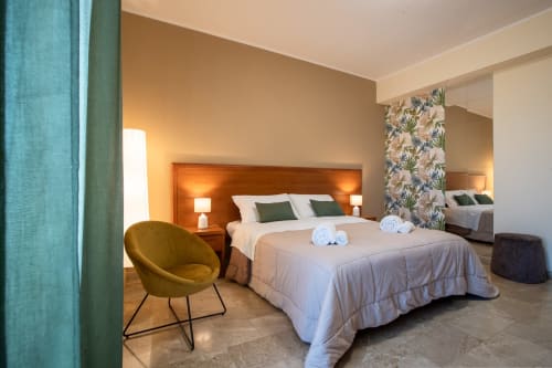 Camera moderna a La Zagara Rooms a Milazzo con testata in legno, biancheria bianca, poltrona verde e parete con decorazioni floreali.