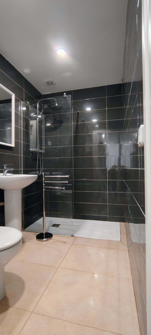 Baño moderno con ducha tipo walk-in de azulejos negros, lavabo blanco, inodoro y piso beige en Hostal Porto Mar, Salou
