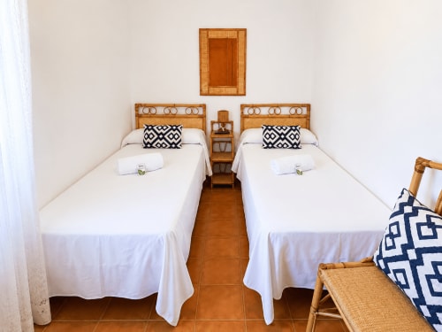 Dos camas en villa Vila Mar de Lua, Oia. Marcos de madera con almohadas geométricas, ropa blanca, piso de barro, mesita de noche.