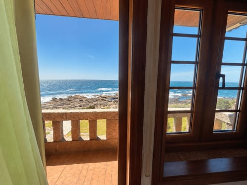 Terraza de villa junto al mar en Oia con vistas a costa rocosa, océano azul y cielo despejado a través de puertas y ventanas de madera
