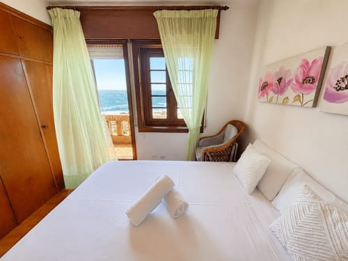 Luminoso dormitorio con vistas al mar en villa de Oia, cama blanca, cuadro de flores rosas y balcón panorámico con vista al océano