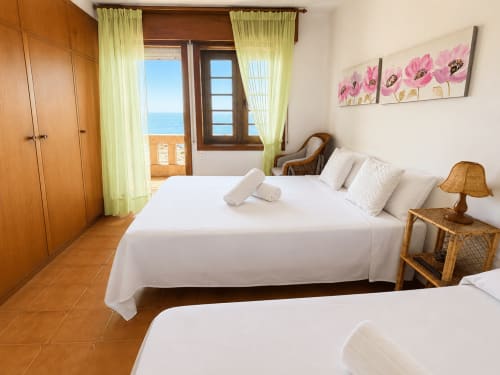 Dormitorio luminoso en Vila Mar de Lua en Oia con vistas al mar, camas blancas, muebles de madera y cortinas amarillas