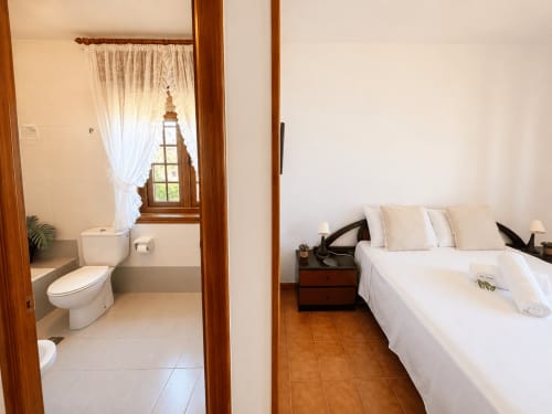 Amplio dormitorio de villa en Oia con vistas al mar, ropa de cama blanca, muebles de madera y baño en suite con ventana