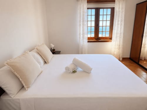 Dormitorio luminoso con cama blanca, almohadas crema, toallas enrolladas y ventana de madera con vistas al mar en villa de Oia