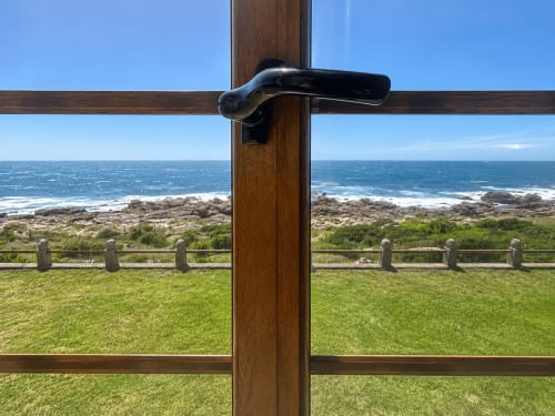 Puerta de madera y vidrio que abre a la terraza de villa en Oia con vistas al océano, acantilados rocosos y jardín verde